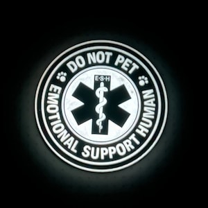 Op de afbeelding: Een ronde sticker in zwart en wit met de tekst "Niet aaien, emotionele ondersteuning mens" en een gestileerd medisch symbool met een slang.