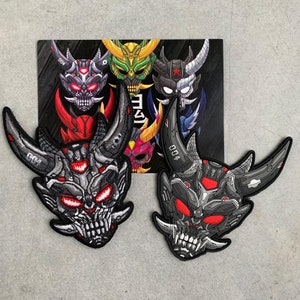 May include: Two embroidered patches featuring a demonic oni mask design with horns, sharp teeth, and glowing red eyes. The patches are primarily black, gray, and red, with intricate detailing. A card with similar designs is in the background.
