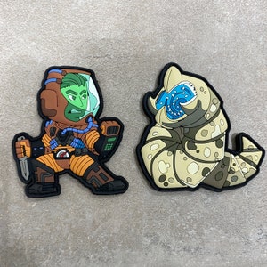 May include: Two embroidered patches, one depicting a green-skinned astronaut with a knife and the other a beige and brown alien creature with a blue mouth.