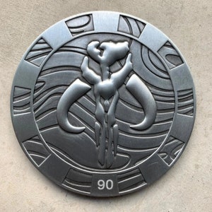May include: A circular, silver-colored metal emblem featuring a stylized skull with horns and a central figure. The emblem has a textured surface with engraved details and the number "90" at the bottom. The design is reminiscent of a Mandalorian symbol.