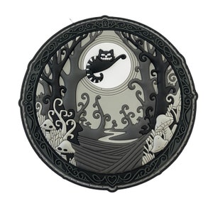 Puede incluir: Un diseño circular en negro, gris y blanco con el Gato de Cheshire. La imagen muestra al gato sonriendo frente a una luna llena, ambientado en una escena de bosque con árboles, setas y un camino. El diseño está enmarcado por un borde decorativo.