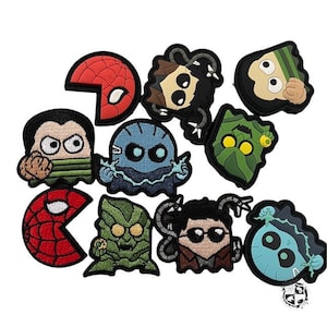 May include: A collection of colorful, cartoon-style patches featuring various characters. The patches include a red Spider-Man mask, a blue ghost, and other figures with distinct facial expressions and designs. The patches are likely for clothing or accessories.