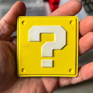 Könnte beinhalten: Ein gelbes Quadrat mit einem weißen Fragezeichen in der Mitte. Das Quadrat hat ein kleines Loch in der oberen rechten Ecke.