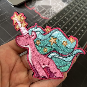 May include: Embroidered patch of a pink unicorn with a white horn and teal mane decorated with gold stars. The patch has a pink border and a cartoon design, suitable for attaching to clothing or accessories.