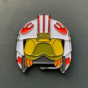 May include: An enamel pin of a Star Wars X-wing pilot helmet. The pin shows a white helmet with red and orange details, a yellow visor, and a black microphone. The design is highly detailed.