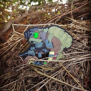 Könnte beinhalten: Eine grüne, braune und blaue Comicfigur mit Helm und Gewehr. Die Figur hockt in einem Stück trockenem Gras.