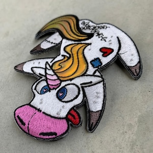 May include: A white unicorn with a pink horn, blue eyes, and a red tongue is stitched together with a red heart and blue bandages. The unicorn has a yellow and orange mane and tail.