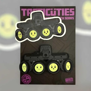 Könnte beinhalten: Zwei Aufkleber mit einem schwarzen Zeichentrick-LKW mit gelben Smiley-Rädern. Der Text "TACTICUTIES" steht oben und "HIWEZ" unten. Die Aufkleber befinden sich auf einem dunklen Hintergrund.