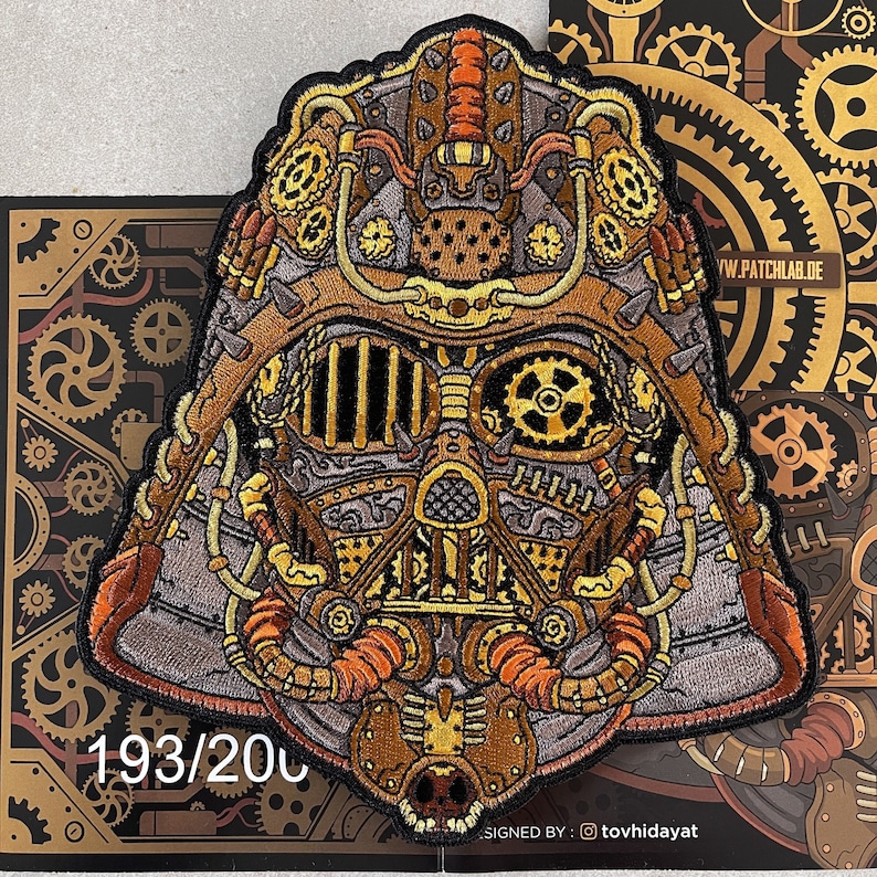May include: A detailed embroidered patch of a steampunk-style Darth Vader helmet with intricate gears and pipes. The patch is brown, gold, and silver with a black background. The patch is numbered 193/200.