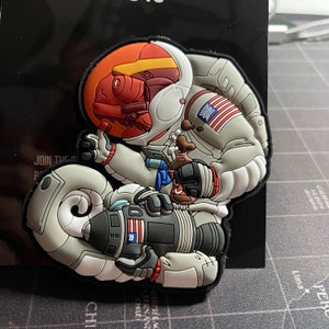 May include: A white astronaut with a red and orange helmet is riding a rocket. The astronaut is wearing a blue and white striped shirt and a red, white, and blue American flag patch on the sleeve. The rocket is white with a black engine and a red, white, and blue American flag patch on the side.
