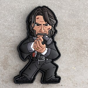May include: Black and white cartoon patch of a man with dark hair, wearing a suit and holding a gun. The man has a serious expression on his face.