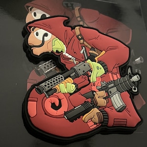 Puede incluir: Un parche de dibujos animados rojo y negro que representa a un personaje con una capa con capucha, sosteniendo múltiples armas de fuego. El personaje tiene un bigote distintivo y guantes verdes. El parche tiene un efecto 3D.