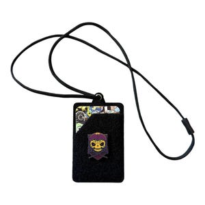 May include: Black felt cardholder with a black lanyard. The cardholder features a colorful pin of a skull with a purple helmet and crossed swords. The cardholder is designed to hold cards and has a pocket.