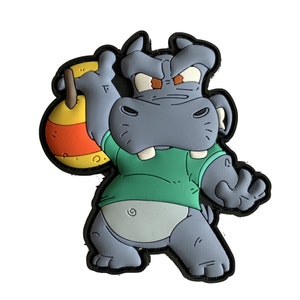 May include: A cartoon hippo patch in shades of gray, green, orange, and yellow. The hippo is wearing a green shirt and holding a round object. The patch has a black border and is set against a white background.