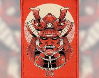 Pode incluir: Patch bordado com uma ilustração detalhada de um capacete e rosto de samurai em tons de vermelho e preto. O desenho é colocado sobre um círculo branco e um fundo retangular vermelho. O patch tem uma borda costurada.
