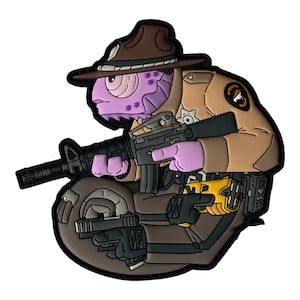 Puede incluir: Un parche de estilo caricatura que representa a un alienígena púrpura con uniforme y sombrero marrón, armado con un rifle y una pistola. El parche tiene un borde negro e incluye una insignia y un cinturón de herramientas.