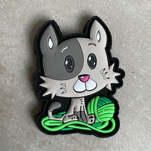 May include: A grey and white cartoon cat with large eyes and a pink nose is sitting on a green ball of yarn. The cat is holding a ball of green yarn in its paws. The cat is surrounded by a green ball of yarn.