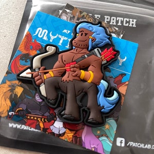 May include: A colourful patch featuring a cartoon centaur with a bow and arrow. The centaur has brown skin, a blue mane, and is holding a bow and arrow with red fletching. The patch is on a blue background with the words "MYTHIC PATCH".