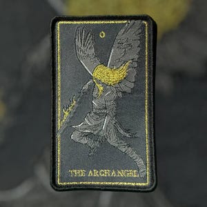 Può includere: Un patch ricamato nero e oro con un angelo con le ali spiegate che tiene una spada. Il testo "THE ARCHANGEL" è ricamato nella parte inferiore del patch.