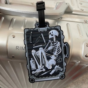 May include: A black luggage tag featuring a skeleton inside a suitcase. The skeleton is holding a bottle and a weapon. The tag has a silver rim with the word "RIM" visible.