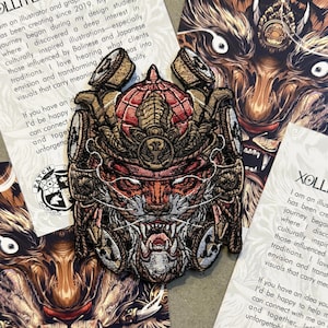 Op de afbeelding: Geborduurde patch met een gedetailleerd ontwerp van een samoeraimasker in rood, goud en bruin. De patch is omgeven door kaarten met illustraties en tekst. De patch heeft een gedetailleerd ontwerp.