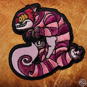May include: Embroidered patch featuring the Cheshire Cat from Alice in Wonderland. The patch has a black border and depicts the cat in shades of pink, purple, and red. The cat is smiling with yellow eyes.