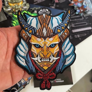 May include: An embroidered patch depicting a samurai warrior's mask in blue, white and gold. The mask has a fierce expression with sharp teeth and a long, pointed nose, and is tied with a red rope around the neck.