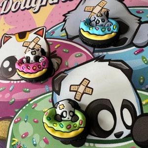 Könnte beinhalten: Drei Comic-Tier-Anhänger mit Pflastern auf dem Kopf. Jeder Anhänger trägt einen Donut mit bunten Streuseln. Die Anhänger sind eine weiße Katze, eine graue Katze und ein schwarz-weißes Panda.