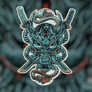 Peut inclure: Un patch brodé représentant un masque oni vert et bleu avec des épées croisées et un motif de corde. Le masque oni a une expression féroce avec des dents pointues et des cornes.