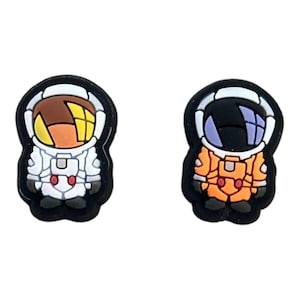 May include: Two astronaut shoe charms, one with a white spacesuit and orange helmet, the other with an orange spacesuit and black helmet.
