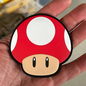 May include: A red and white mushroom-shaped patch with black trim. The mushroom has a white spot on top and two black eyes.
