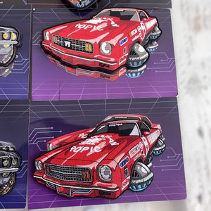 Puede incluir: Parche de coche bordado rojo con la palabra "POP IC" en el capó. El parche presenta un diseño de coche vintage con detalles de carreras y logotipos de patrocinadores. El parche está sobre un fondo morado con un diseño de placa de circuito.
