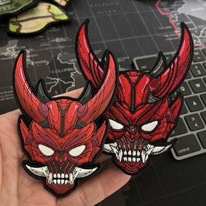 May include: Two red and white embroidered iron-on patches depicting Japanese demon masks with horns. The masks have sharp teeth and white eyes.