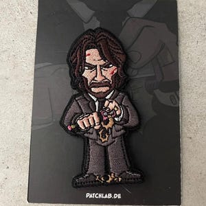 Może przedstawiać: Naszywka haftowana przedstawiająca postać z kreskówki o ciemnych włosach, w czarnym garniturze, z poważnym wyrazem twarzy. Postać trzyma pistolet w jednej ręce, a ołówek w drugiej. Naszywka ma czarne tło i napis "PATCHLAB.DE" na dole.