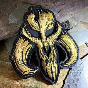 Könnte beinhalten: Ein gold- und schwarz gestickter Aufnäher mit einem stilisierten Schädel mit Hörnern. Der Schädel hat ein detailliertes, strukturiertes Design und ist schwarz umrandet.