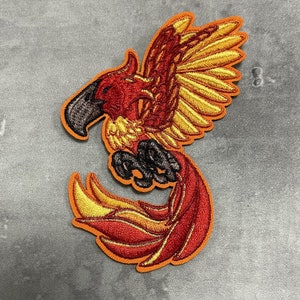 Puede incluir: Parche bordado para planchar de un fénix rojo y amarillo con garras negras y un pico negro. El fénix está en vuelo con las alas extendidas y las plumas de la cola arrastrándose detrás de él.