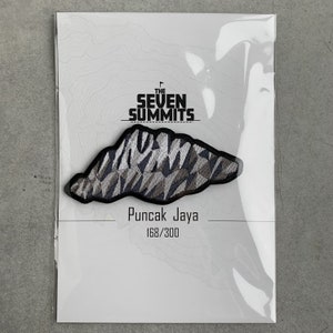 May include: A patch featuring a mountain range design in shades of gray, black, and white. The patch is labeled "Puncak Jaya" with the text "The Seven Summits" above the mountain graphic. The patch is numbered 168/300.