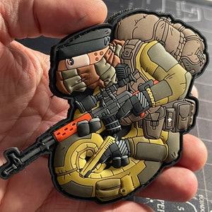 Könnte beinhalten: Ein brauner und beiger Comic-Soldat mit Rucksack und Gewehr. Der Soldat trägt einen braunen Schal und einen schwarzen Hut. Der Soldat hält ein Gewehr in der rechten Hand und eine Pistole in der linken Hand. Der Soldat steht auf einem gelben und braunen Objekt.