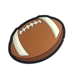 Op de afbeelding: Een bruine, voetbalvormige bedel met witte strepen en stiksels. De bedel heeft een zwarte rand en is ontworpen om op een klassieke American football te lijken.