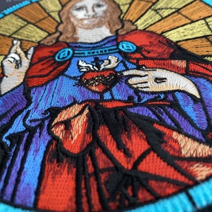 Könnte beinhalten: Gestickter Patch, der ein Buntglasbild von Jesus Christus mit einem Herzen in seiner Brust zeigt. Das Herz ist von Dornen umgeben und rot, während der Hintergrund blau, lila und orange ist.