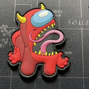May include: A red and black Among Us character with a large, open mouth and a pink tongue sticking out. The character has a blue visor and yellow teeth.