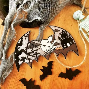May include: A Halloween-themed bat-shaped applique with a haunted house, pumpkins, and graveyard design. The applique is black, gray, and brown, with a white skull in the center. Three black bat silhouettes are scattered below. A gray fabric and a skeleton are in the background.