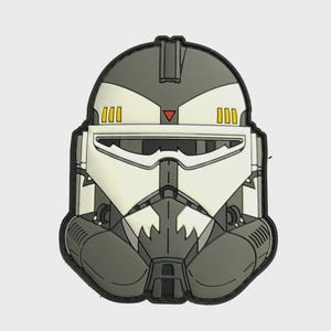 Pode incluir: Um patch com uma ilustração detalhada de um capacete branco, cinza e preto. O capacete tem um design distinto com uma viseira, um triângulo vermelho e três retângulos amarelos em cada lado. O patch é delineado em preto.