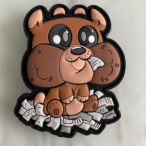 May include: A brown cartoon dog patch with large black eyes and a tan muzzle, holding a piece of paper in its mouth. The dog is sitting on a pile of white and grey paper, with a black border.