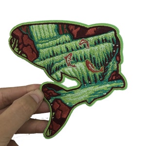 Op de afbeelding: Geborduurde patch met een gestileerd visontwerp met een waterval en koi-karper. De patch heeft een groene rand, met groene, bruine en rode details. De patch wordt door een hand vastgehouden.