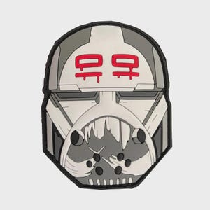 Pode incluir: Um patch com um design de capacete estilizado em tons de cinza, branco e preto. O capacete tem símbolos vermelhos no topo e um design de boca detalhado. O patch tem uma borda preta.