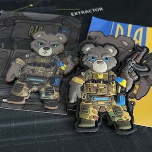 Könnte beinhalten: Zwei gestickte Patches mit Comic-Teddybären, die militärische Ausrüstung tragen. Die Bären halten Waffen und haben die ukrainische Flagge an ihren Armen. Der Text "RIA" befindet sich auf den Westen der Bären. Die Patches befinden sich auf einer schwarzen Oberfläche.