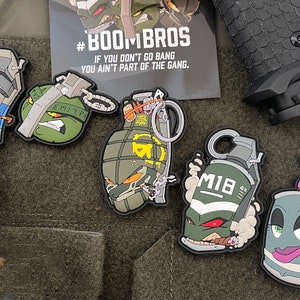 Puede incluir: Cinco parches de granada de dibujos animados coloridos con diferentes expresiones. Los parches están hechos de goma y tienen un efecto 3D. Los parches presentan un esquema de color verde, azul y gris. Los parches tienen una variedad de detalles, incluyendo un lazo rosa, un cigarro y una cadena. Los parches tienen el texto "#BOOMBROS" y "M1B" en ellos.