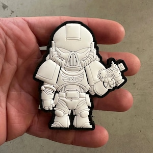 May include: A white and black cartoon-style patch featuring a futuristic soldier with a helmet, armor, and a weapon. The patch has a black border and is held in a hand.
