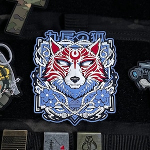 Puede incluir: Parche con un diseño de zorro estilizado en rojo, blanco y azul, con texto japonés. El diseño incluye elementos florales y geométricos. El parche tiene un borde blanco.